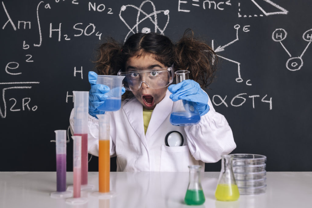 surprised scientist girl with gloves and goggles in lab coat with chemical flasks, blackboard background with science formulas, explosion in the laboratory, back to school concept