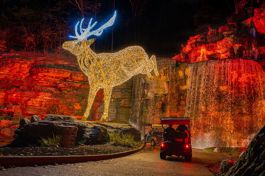 A large buck made of yellow and white lights bounding over a trail with a golf cart traveling under it. In the background is a waterfall flowing over a rock ledge surrounded by red colored lights.