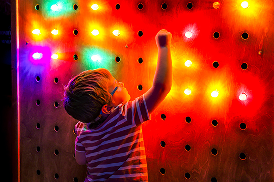 A young boy reaches up to screw in a light bulb on a wall illuminated by bright colorful lights.