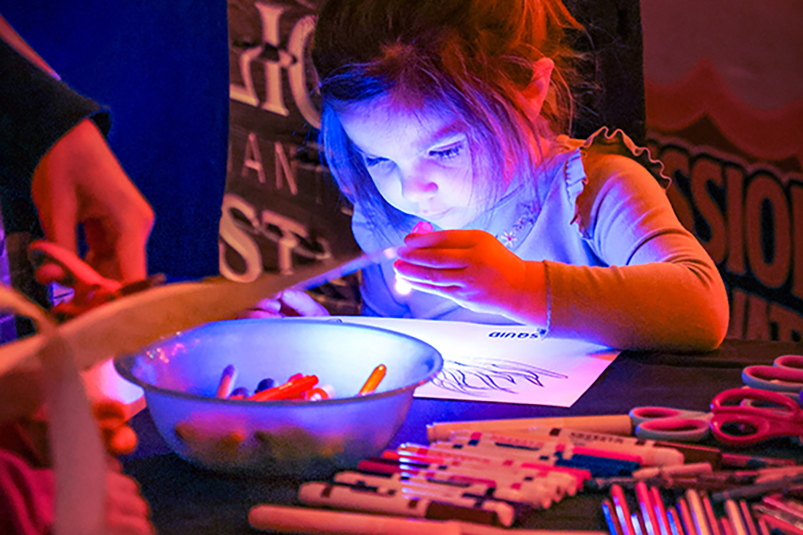 A young girl uses a flashlight to light up her coloring page as she colors with markers.