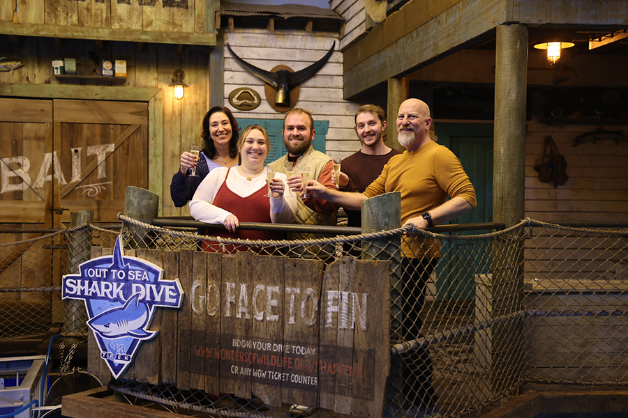 A group of people stand cheers drinks while standing behind a railing that has a "Out To Sea Shark Dive" sign on it.