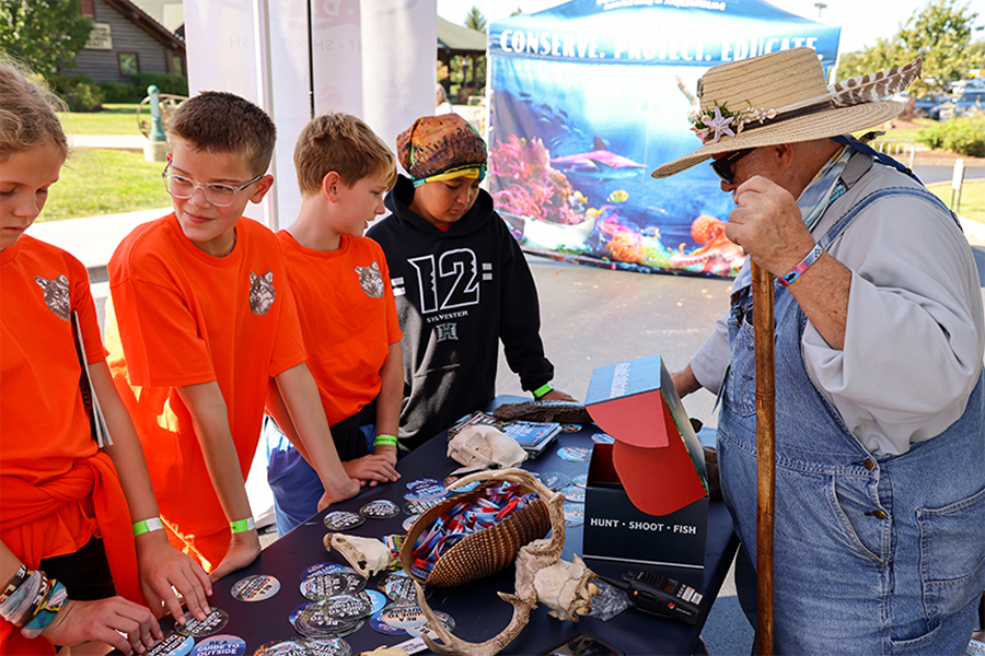 A WOW volunteer shows a group of young boys several artifacts on a table at an outdoor event.