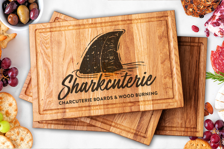 A white background with a variety of fruits, cheeses, and crackers and a stack of charcuterie boards with a wood burned "Sharkcuterie" logo.