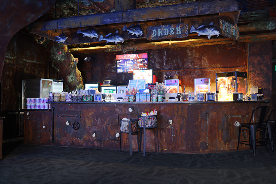 A small counter wraps around a snack bar with all manner of snacks and candies on the counter, a popcorn and pretzel machine in the background and a digital menu board on the back wall.