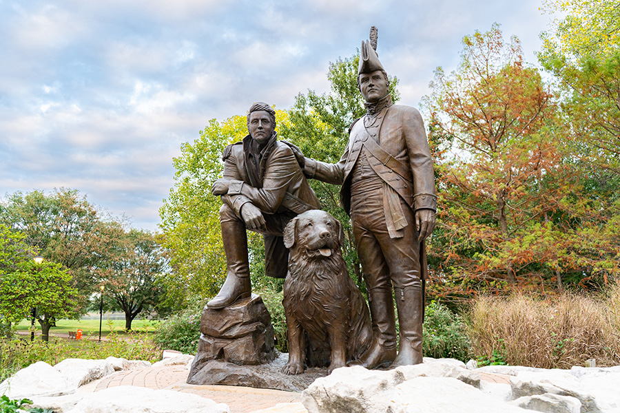 A statue of Lewis and Clark with their faithful dog stand perched on a rock in front of a treeline.
