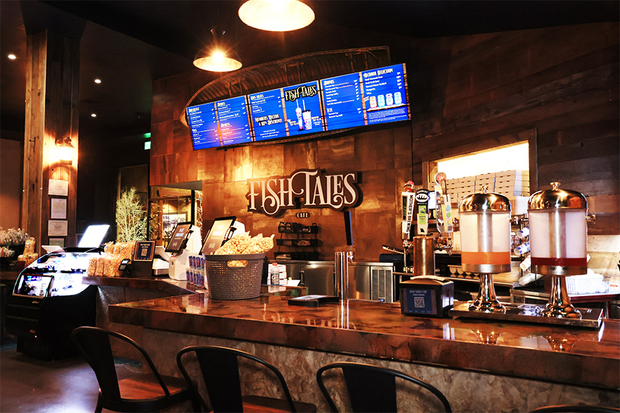 A hard counter top full of snacks and merchandise with a digital menu board above on the wall directly above text that says: "Fish Tales Cafe".
