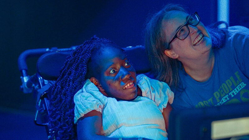 A young African-American girl with Cerebral Palsy smiles with her adoptive mother smiling beside her.