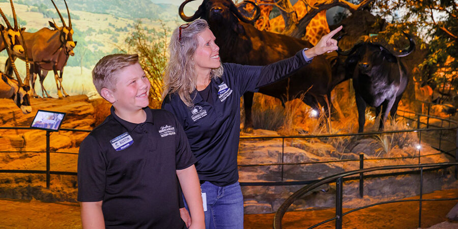 A volunteering mom smiles and points at an exhibit while her volunteer son smiles.