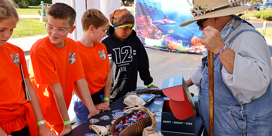A WOW volunteer shows a group of young boys several artifacts on a table at an outdoor event.